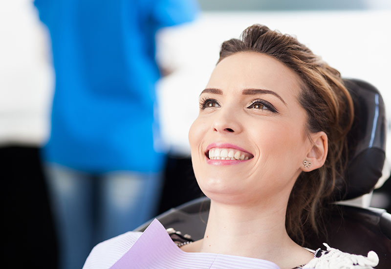 Eine junge Frau mit schwarzen Haaren sitzt lächelnd auf einem Zahnarztstuhl.