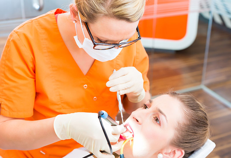 Eine Zahnärztin im orangenen Kittel führt bei einer Frau eine Zahnreinigung durch.